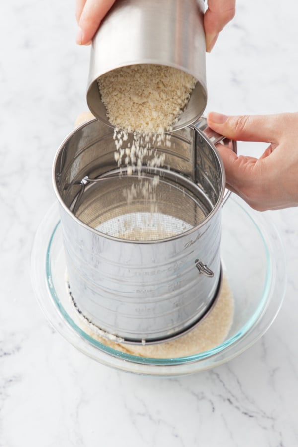 Pouring more ground almonds into a metal flour sifter.