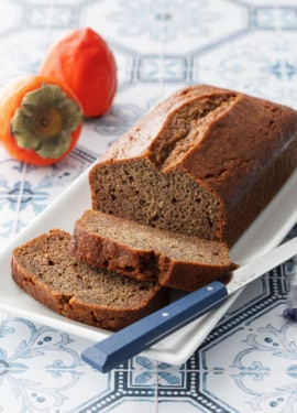 Loaf of Spiced Persimmon Bread on a white cake plate, two slices cut and laying down, with two hachiya persimmons in the background.