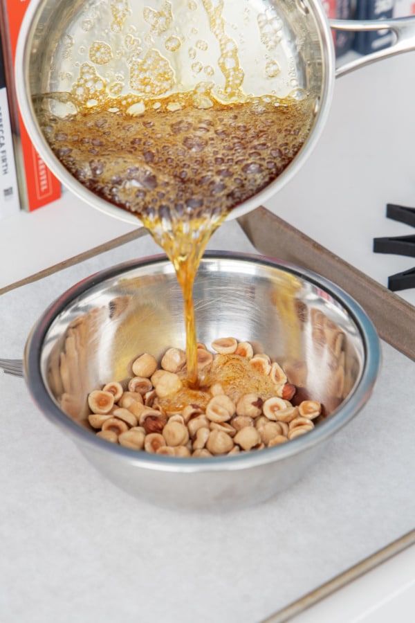 Pouring half of the hot liquid caramel over coarsely chopped hazelnuts in a bowl.