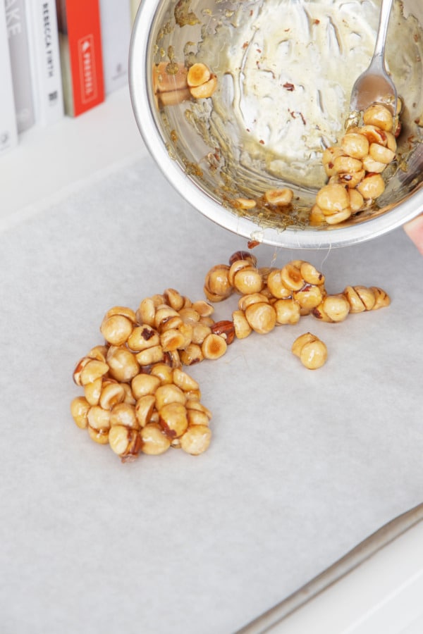 Pouring caramel-coated chopped hazelnuts onto a parchment-lined baking sheet.