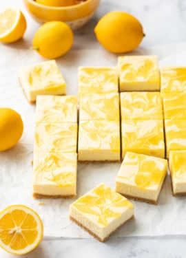Squares of Meyer Lemon Cheesecake Bars with a swirl of lemon curd on top, on a piece of parchment with a few Meyer lemons scattered around.