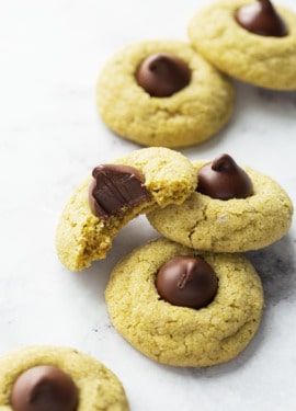 Scattered Pistachio Butter Blossom Cookies on a white background, one cookie with a bite out of it to show the dark chocolate and soft texture of the cookie.