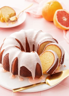 Grapefruit Olive Oil Bundt Cake with two cut slices leaning against each other, on a white cake plate and cut grapefruit and a slice of cake in the background.
