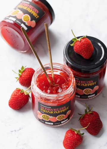 Three jars of homemade Strawberry Passionfruit Jam, one jar open with two gold spoons stuck inside, and a few fresh strawberries scattered around.