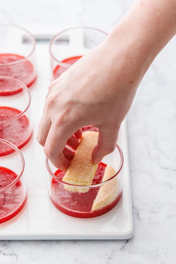 Layering cut ladyfinger cookies in clear ramekins on top of a layer of strawberry sauce.