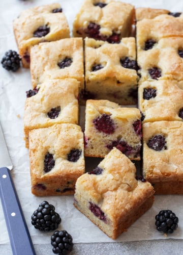 Cut squares of Blackberry Butter Cake on a piece of parchment, with a small blue knife and a few fresh blackberries scattered around.