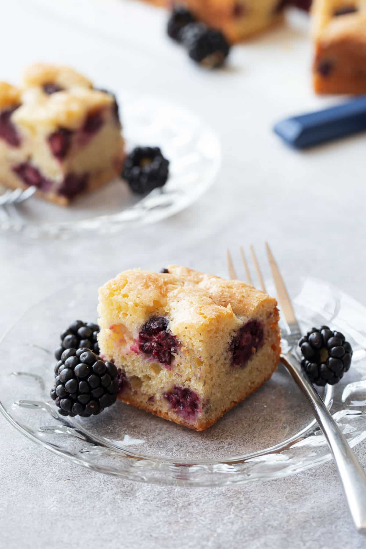 Cut square of Blackberry Butter Cake on a glass dessert plate, with silver fork and fresh blackberries.