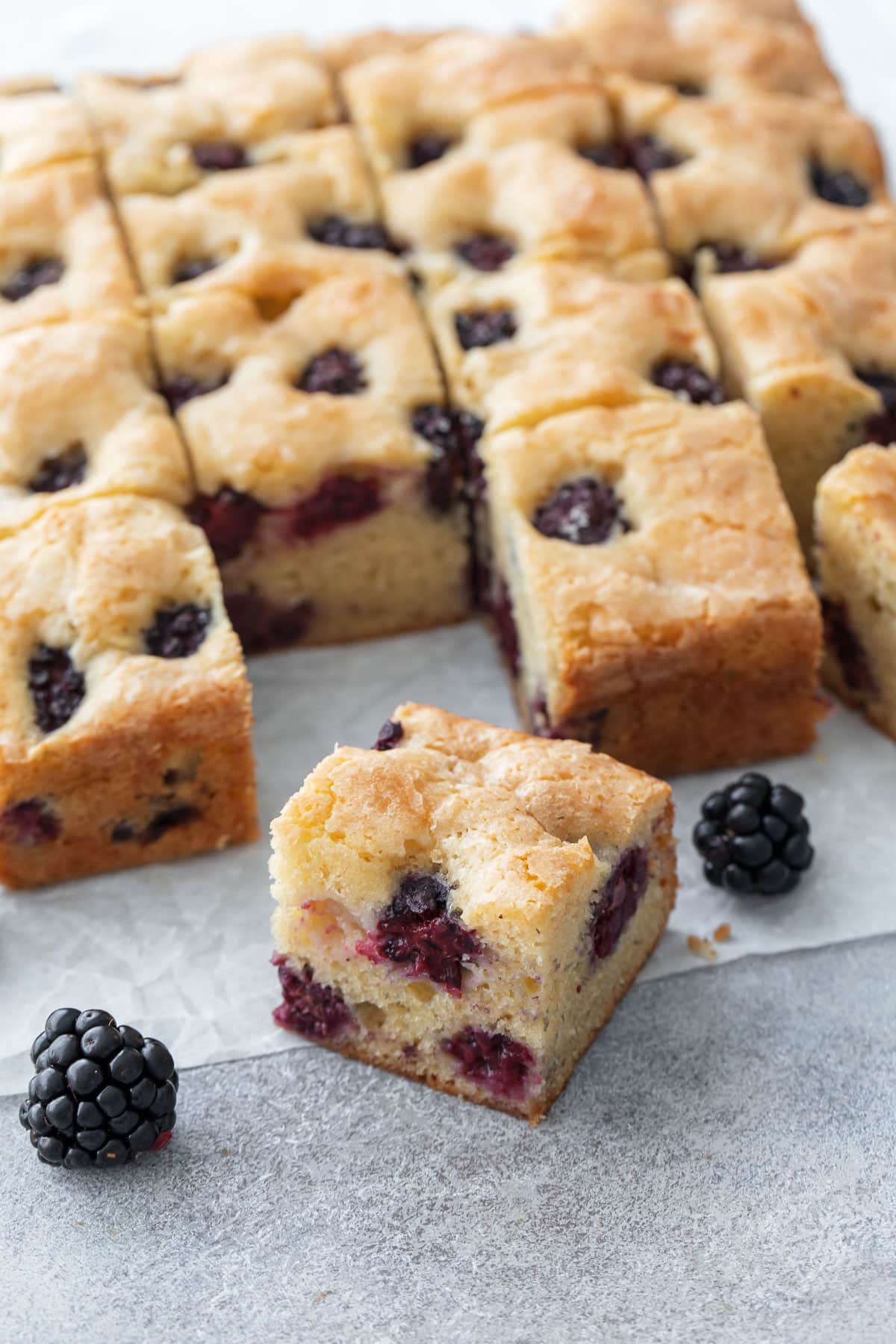 Blackberry Butter Cake cut into squares and sitting on a piece of parchment, with fresh blackberries scattered around.