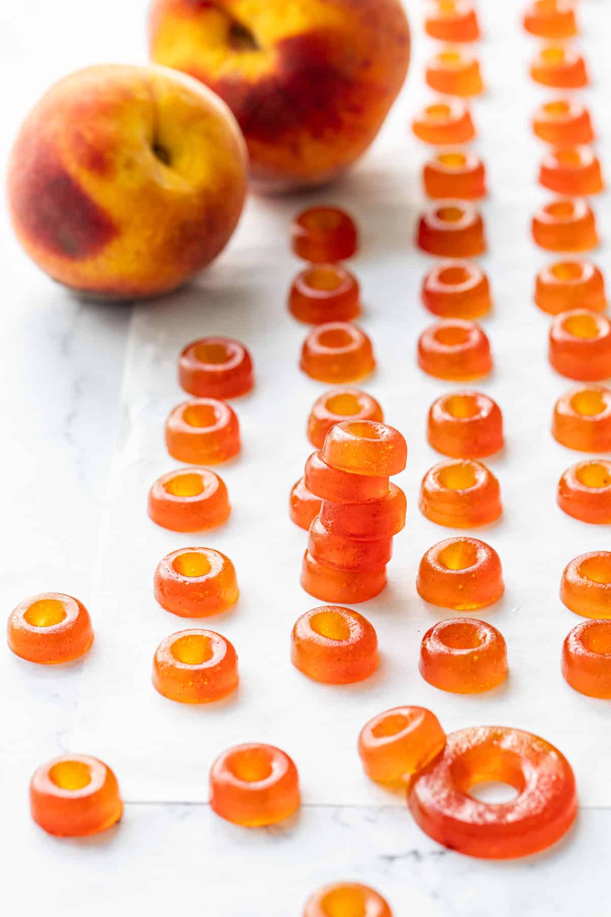 Lines of Homemade Gummy Peach Rings on a piece of parchment, with two fresh peaches in the background.
