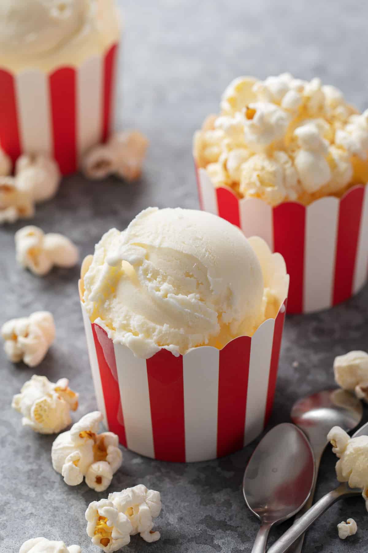 Three red and white striped containers with scoops of Buttered Popcorn Ice Cream, on a gray background with popped popcorn scattered around.