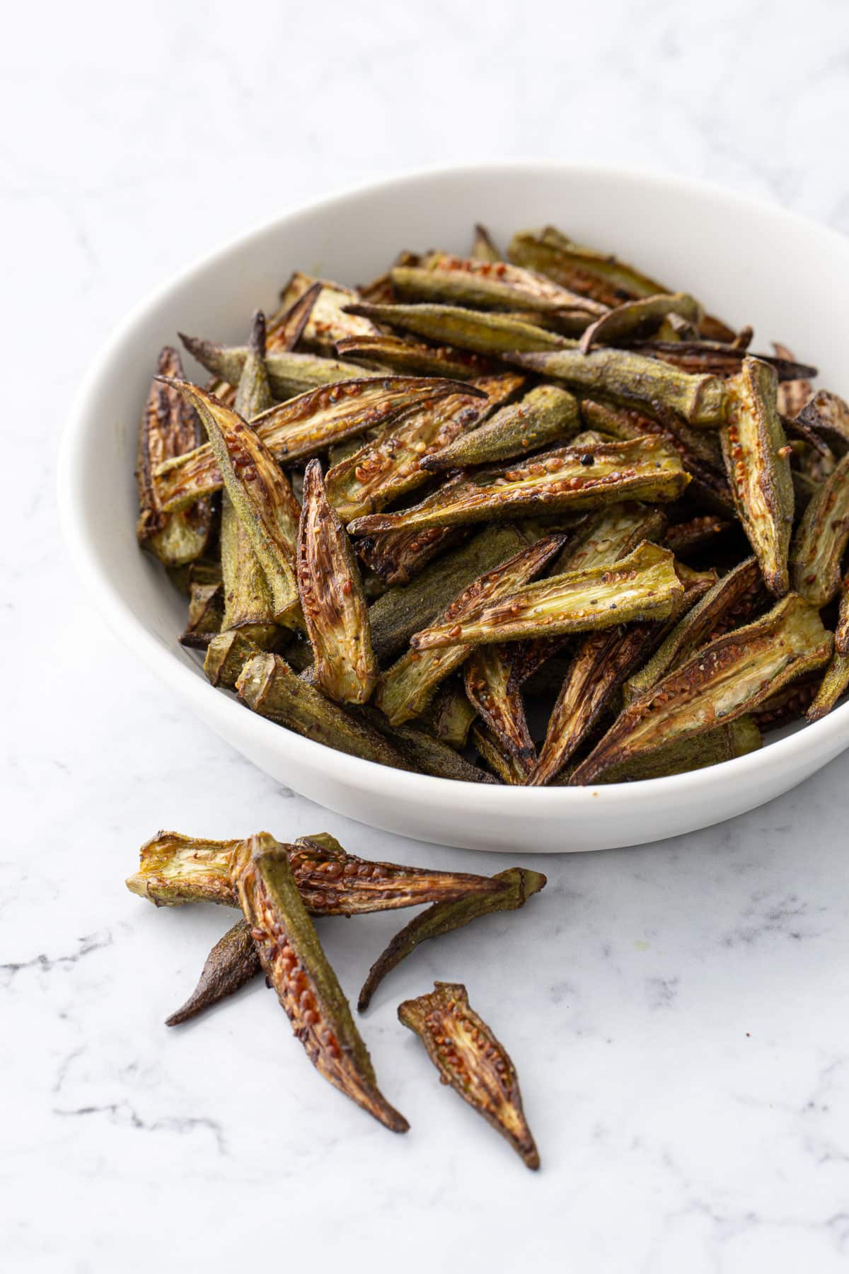 White bowl piled with Crispy Oven-Roasted Okra, a few laying on the side on a marble countertop.