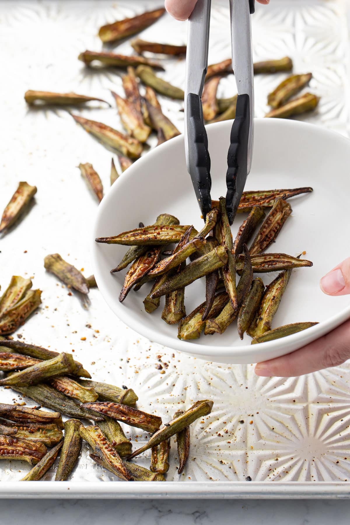 Tongs transferring Crispy Oven-Roasted Okra from a sheet pan to a serving bowl.