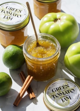 Jars of Green Tomato Jam on a marble background with whole green tomatoes, limes, and cinnamon sticks surrounding an open jar.