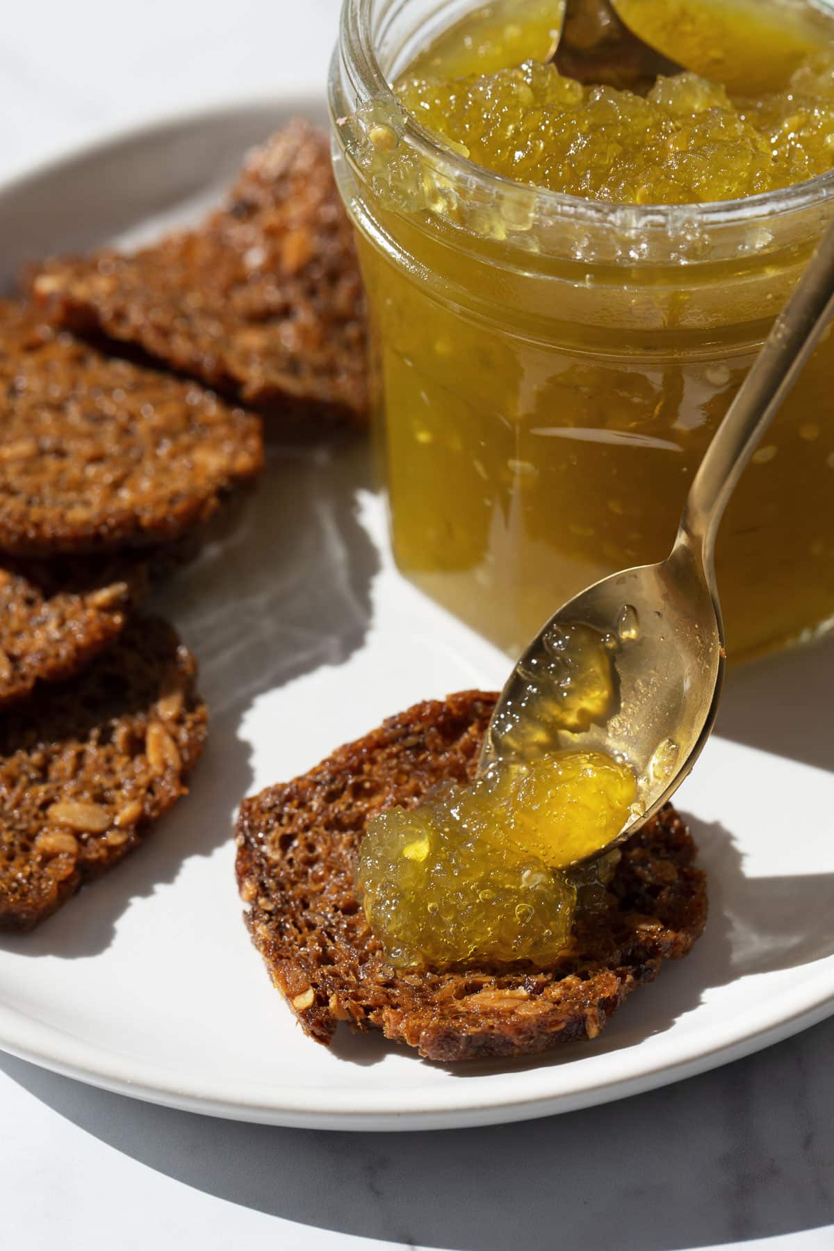 Spoon dropping a dollop of Green Tomato Jam on a rye cracker, on a plate with more crackers and an open jar of jam in the background.