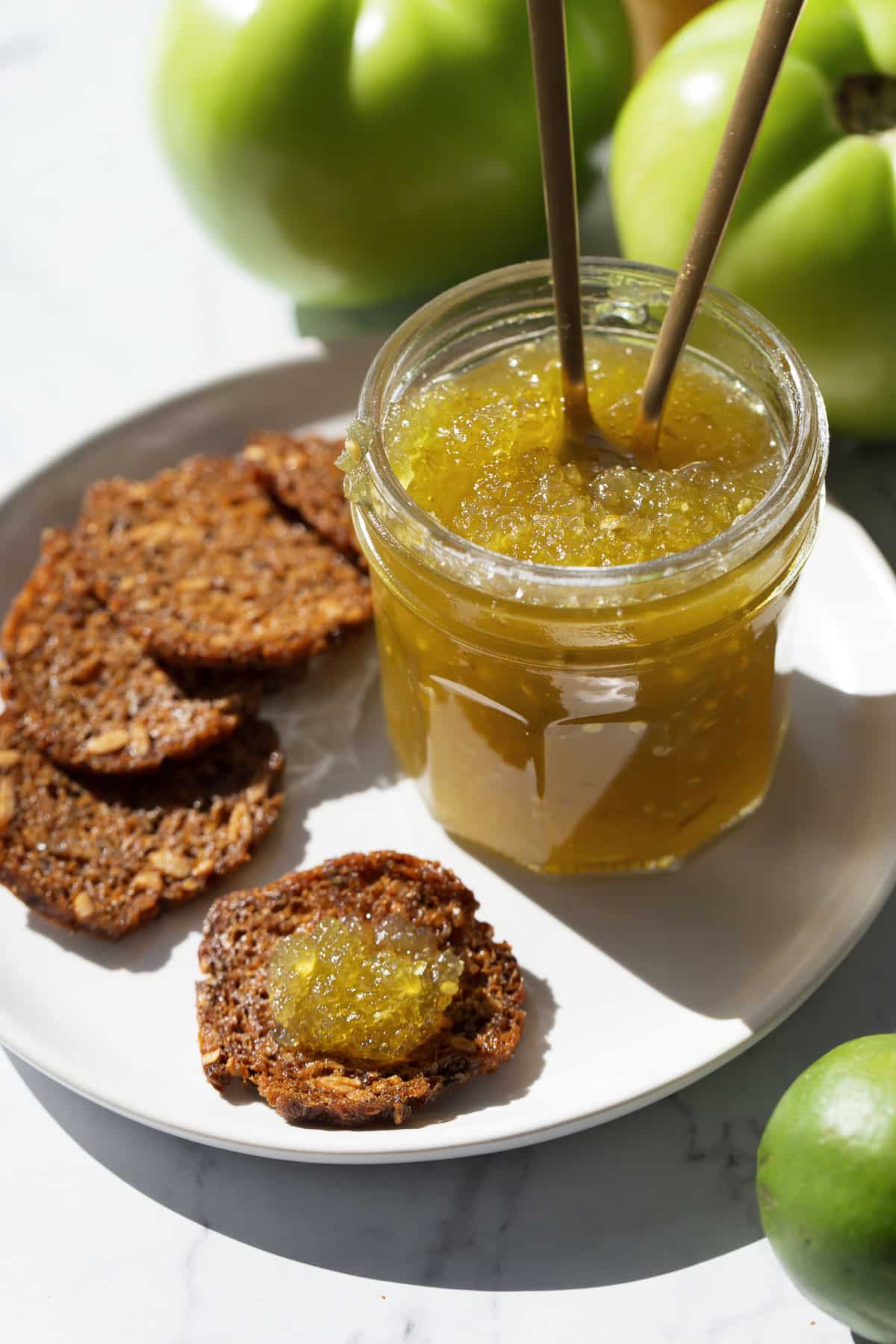 Open jar of Green Tomato Jam with two spoons, on a plate with crackers and bright green tomatoes and limes in the background.