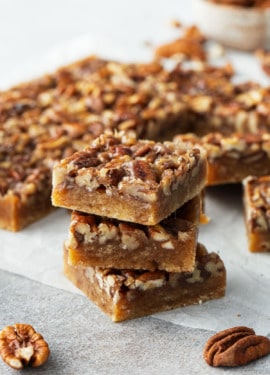 Stack of three square cut Pecan Pie Shortbread Cookie Bars on parchment, with the rest of the pan of bars in the background.