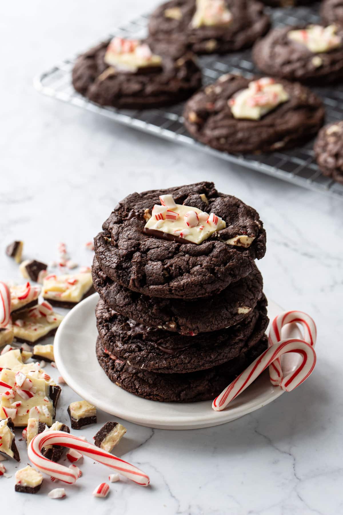 Stack of Chocolate Peppermint Bark Cookies on a small plate, with candy canes and broken up peppermint bark scattered around, rack of cookies cooling in the background.
