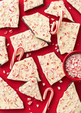 Overhead view of rough broken pieces of homemade peppermint bark scattered on a red background with bits of crushed candy canes scattered around.