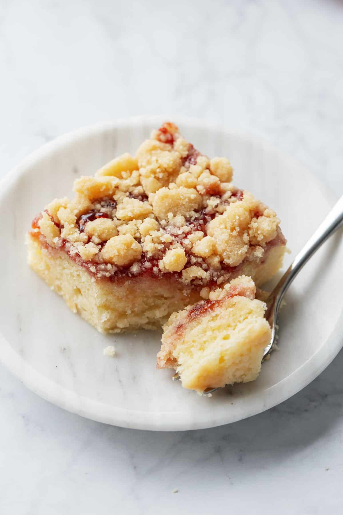 Square of Jammy Sour Cream Crumb Cake on a marble dessert plate with a bite sitting on the fork showing the tender texture of the cake.