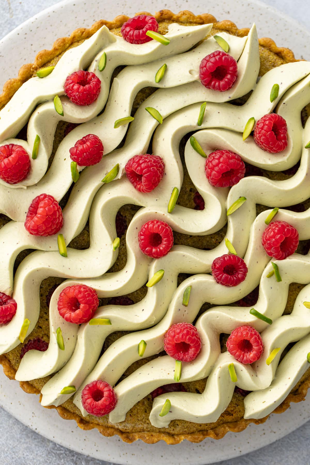 Closeup of the top of a pistachio raspberry tart, piped whipped ganache in flowing waves and garnished with fresh raspberries and slivered pistachios.
