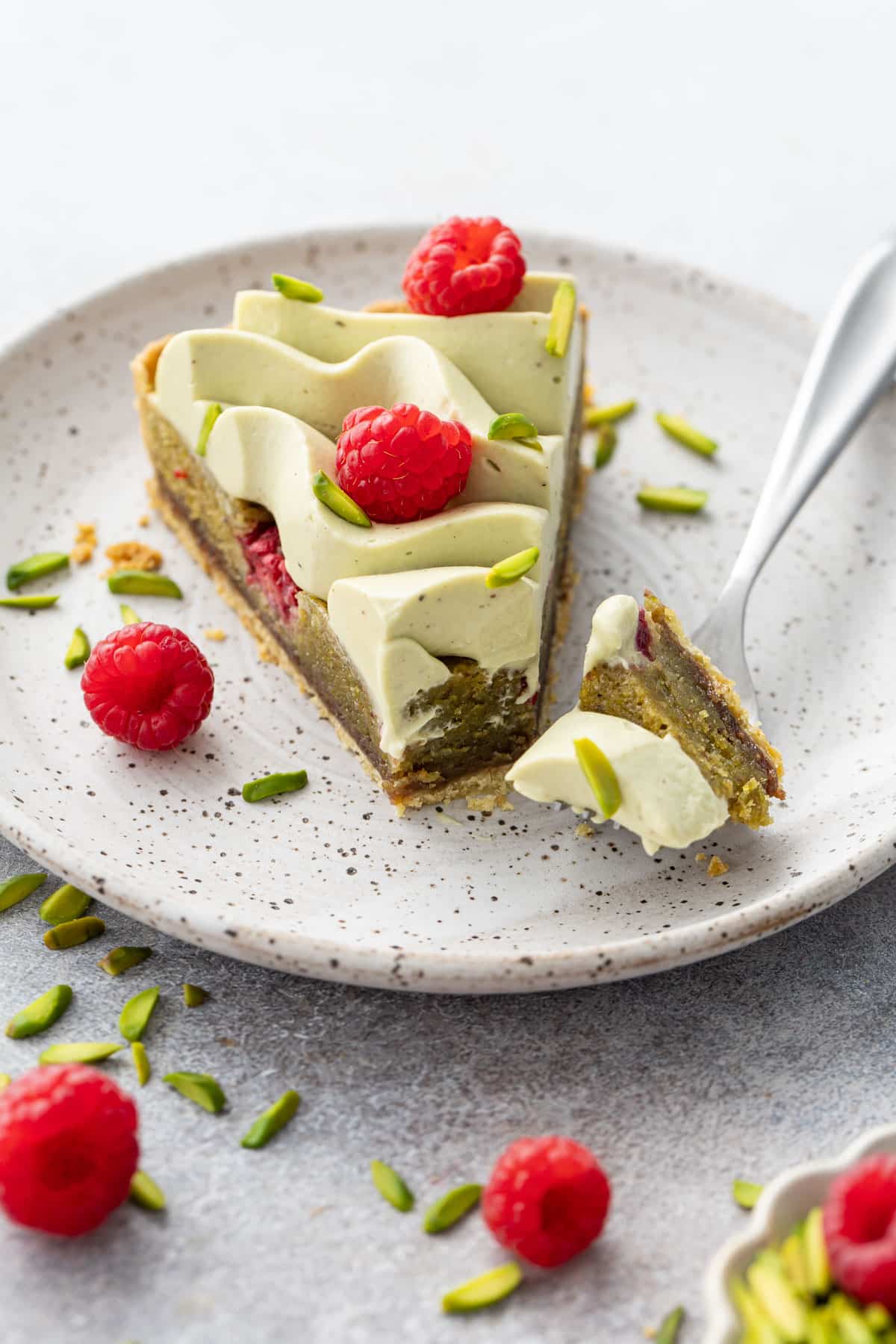 Slice of Raspberry & Pistachio Frangipane Tart with Whipped Pistachio Ganache on a speckled plate, with a fork and a bite to show the consistency of the inside.