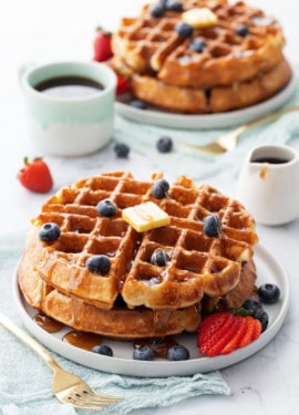 Plates with stacks of golden brown sourdough Belgian waffles topped with berries, a pat of butter and drizzled syrup.