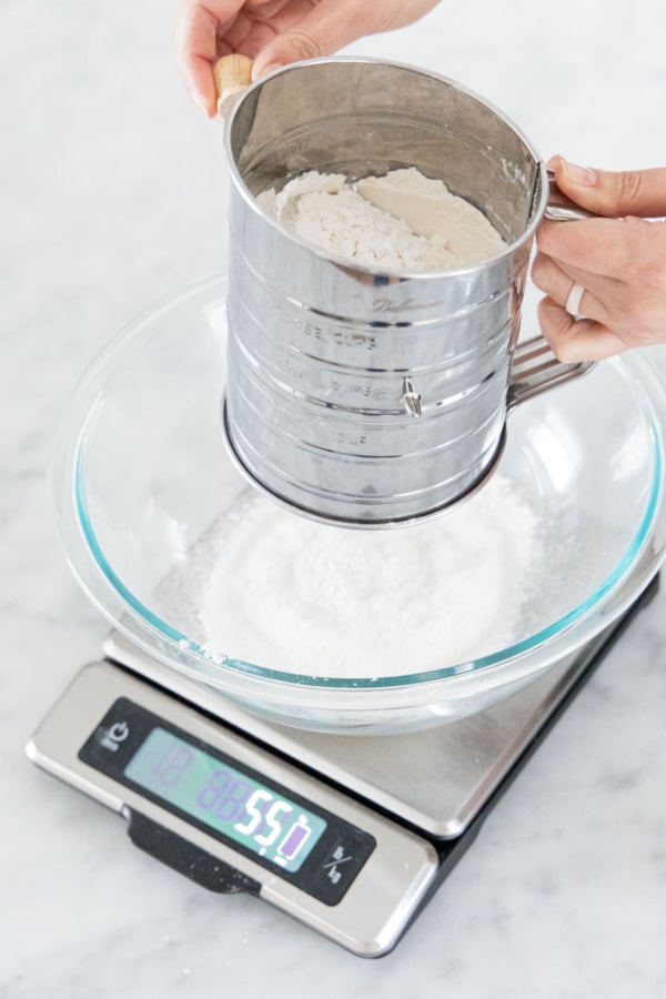Sifting flour into a glass mixing bowl sitting on a digital scale.