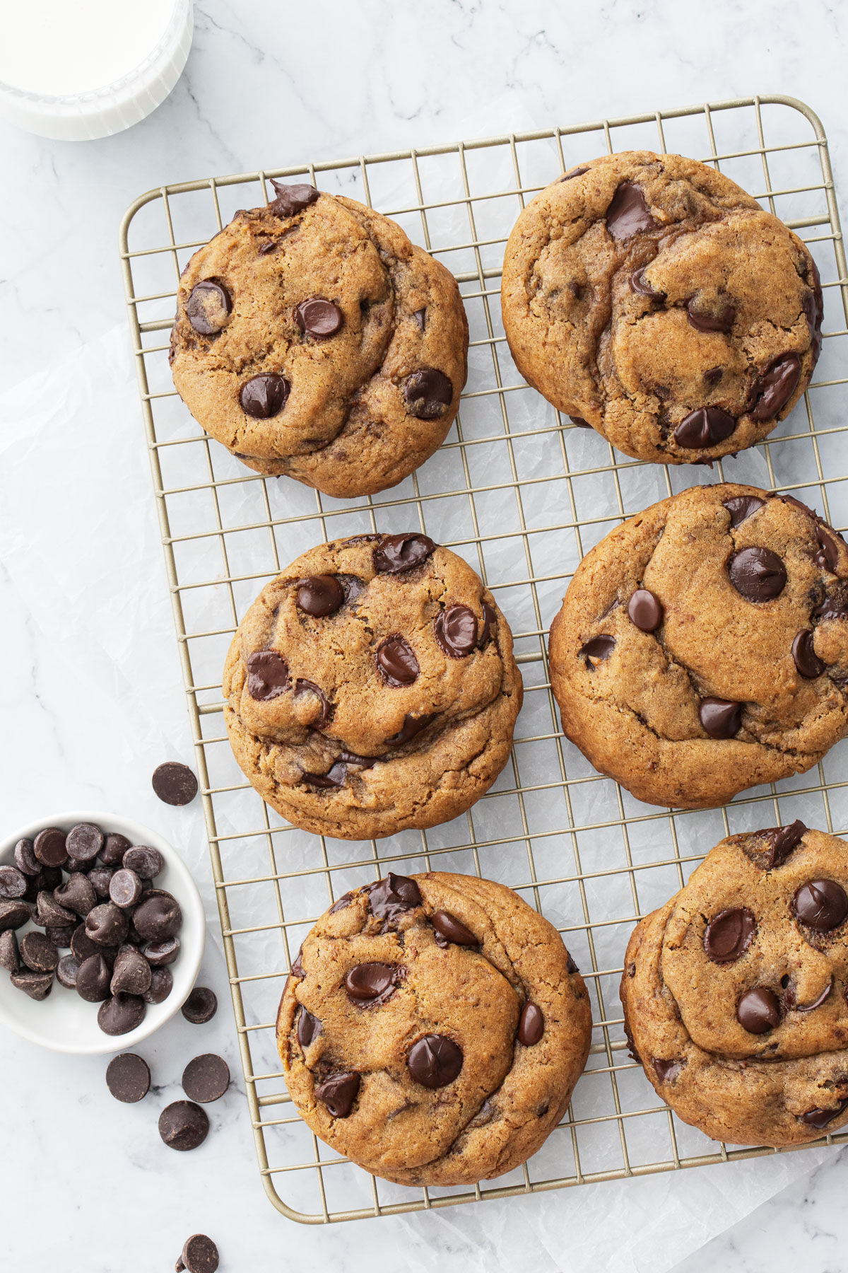 Overhead view of six extra-large Brown Butter Chocolate Chip Cookies on a wire rack with a glass of milk and bowl of chocolate chips on the side.