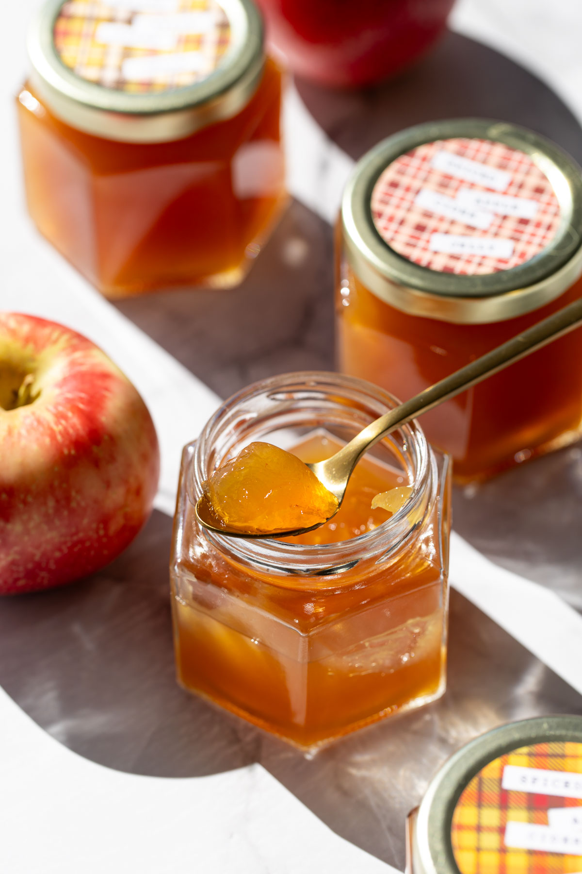 Jar of Spiced Apple Cider Jelly with a spoonful of jelly resting on top to show the glass-like appearance and texture, with more jars and a few apples in the background.