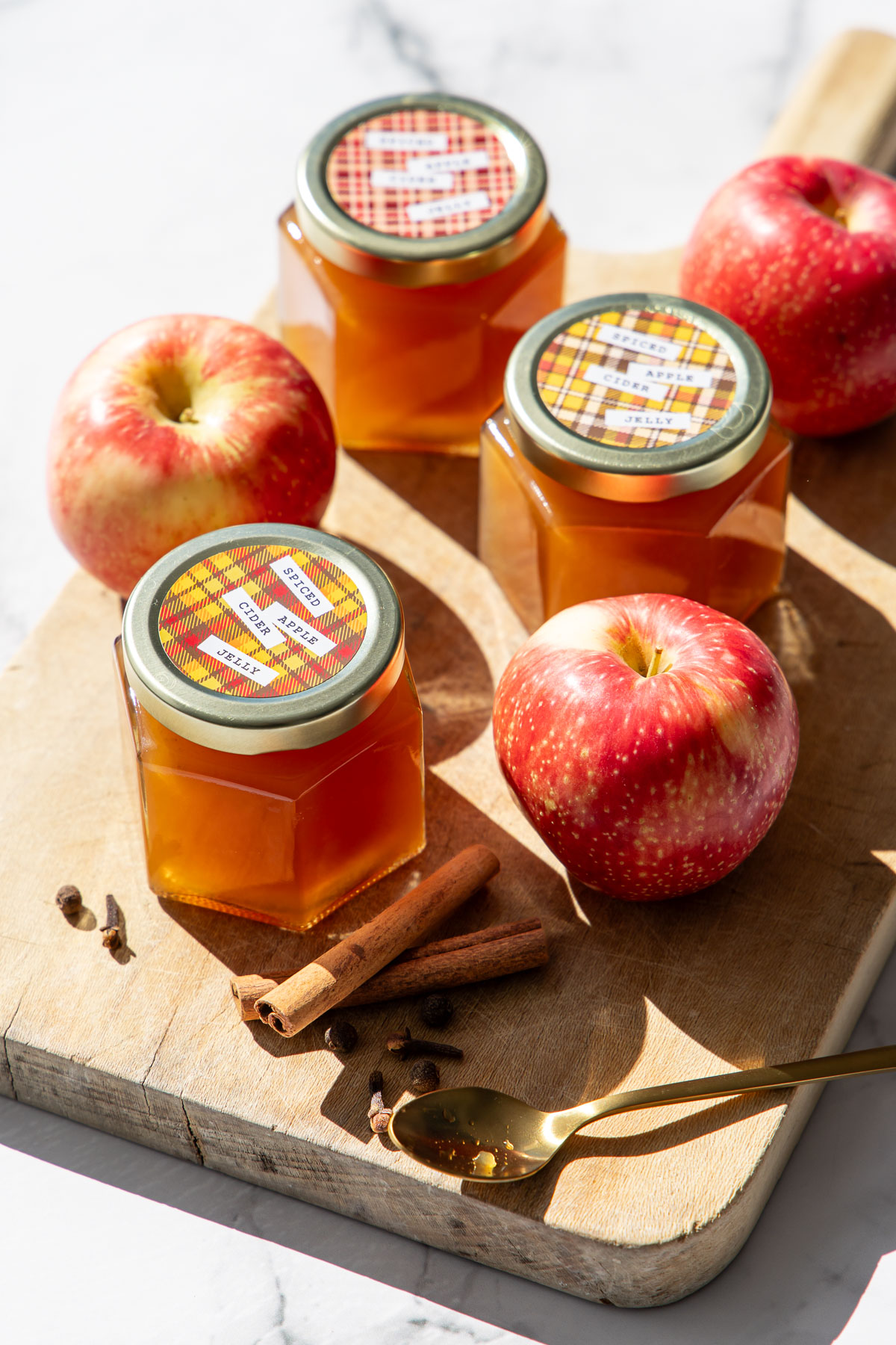 Three jars of Spiced Apple Cider Jelly with designed printable labels, on a wooden board with fresh apples and whole spices.