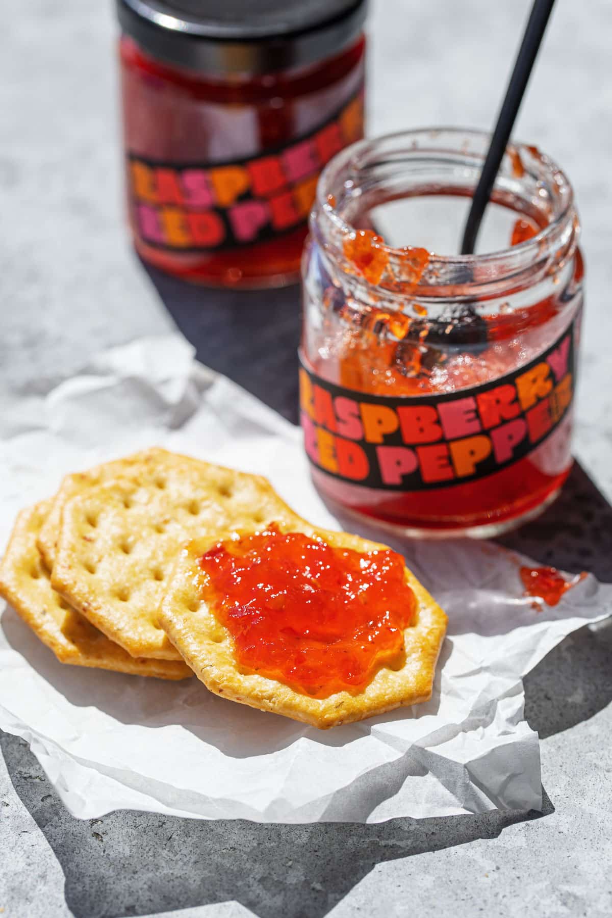 Stack of crackers, top one spread with Raspberry Red Pepper Jelly, with an open jar of jelly with a spoon in the background.