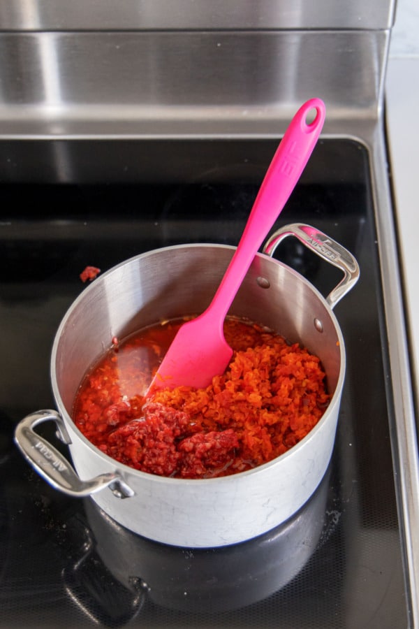 Chopped peppers and red wine vinegar in a stainless saucepan with pink spatula.